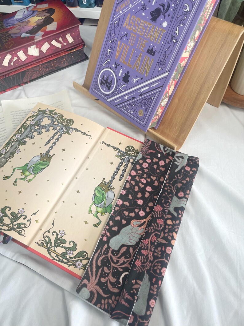 K&ouml;nnte beinhalten: Ein Hardcover-Buch mit dem Titel "Assistant to the Villain" in einem h&ouml;lzernen Buchst&auml;nder. Der Einband ist lila mit goldfarbenen Buchstaben und dekorativen Elementen. Offene Buchseiten zeigen Illustrationen von Fr&ouml;schen. Ein floraler Lesezeichen ist ebenfalls sichtbar.