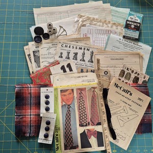 May include: A collection of vintage sewing and craft supplies, including buttons, fabric scraps, sewing patterns, and paper ephemera. The items are arranged on a green grid surface, suggesting a crafting or sewing project.