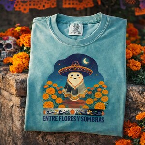 May include: A teal t-shirt featuring a graphic of a ghost wearing a sombrero, surrounded by orange marigold flowers under a crescent moon. The text "ENTRE FLORES Y SOMBRAS" is printed below the illustration. The shirt has a Comfort Colors label.