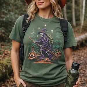 May include: An olive green t-shirt featuring a graphic print of a skeleton dressed as a witch in a purple robe and hat, carrying a broomstick and a backpack. The scene includes pine trees, carved jack-o'-lanterns, and small ghost figures.