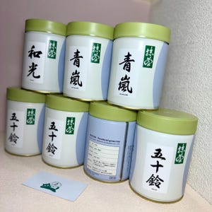 May include: A selection of matcha powdered green tea tins. The tins are white with a light blue stripe and a green lid. Japanese characters are printed on the tins. A business card with a green logo is in the foreground.