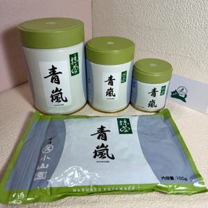 May include: Three white and green containers of varying sizes, labeled with Japanese text. A rectangular package with Japanese text and the brand name "MARUKYU KOYAMAEN" is in front of the containers. A small card with a cat illustration is on the right.