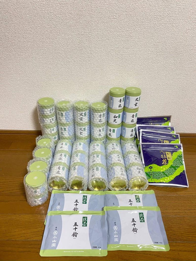 May include: An assortment of packaged Japanese matcha tea products. The image shows several cylindrical containers and rectangular packets, all with Japanese text and green and white packaging. The products are arranged on a light-colored surface.
