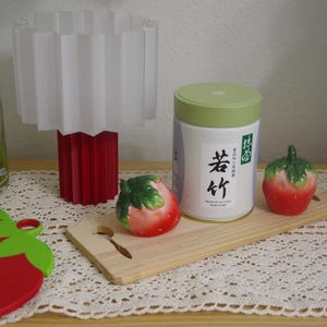 May include: A white and red lamp with a pleated shade sits next to a white tea canister with Japanese text and two ceramic strawberry figurines. A red strawberry-shaped cutting board is in the foreground.