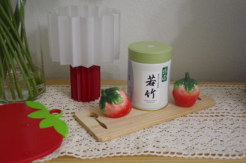 May include: A white and red lamp with a pleated shade sits next to a white tea canister with Japanese text and two ceramic strawberry figurines. A red strawberry-shaped cutting board is in the foreground.