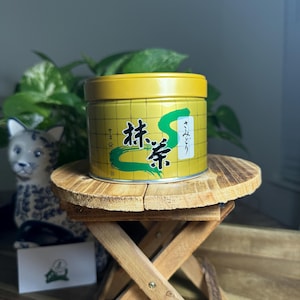 May include: A yellow tin of matcha tea with Japanese text and a green 'S' design. The tin sits on a small wooden stool, with a ceramic cat figurine and green plant in the background.