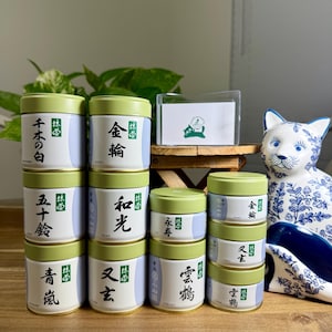 May include: A collection of cylindrical metal tea canisters with light green lids and white bodies featuring black Japanese calligraphy and small green labels. A white card with a green cat logo and a blue and white ceramic cat figurine are present.