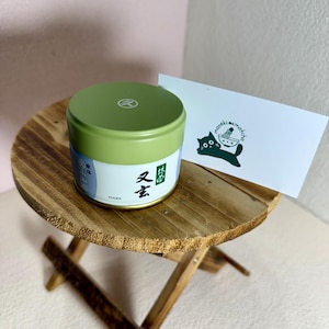May include: A green and white tin of matcha tea with Japanese characters and the word "Yugen" on the label. The tin sits on a small wooden table next to a white card with a black cat logo and the words "maneki matcha".