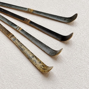 May include: A set of four long, slender bamboo tea scoops, each featuring a curved end. The scoops display natural variations in color, ranging from dark charcoal gray to mottled light brown, with visible bamboo nodes along the stems.