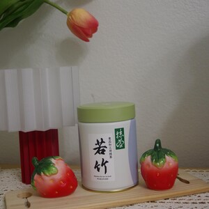 May include: A still-life arrangement featuring a white and green matcha tea tin with Japanese text, flanked by two ceramic strawberry figurines. The scene is set on a wooden board, with tulips and a lamp in the background.