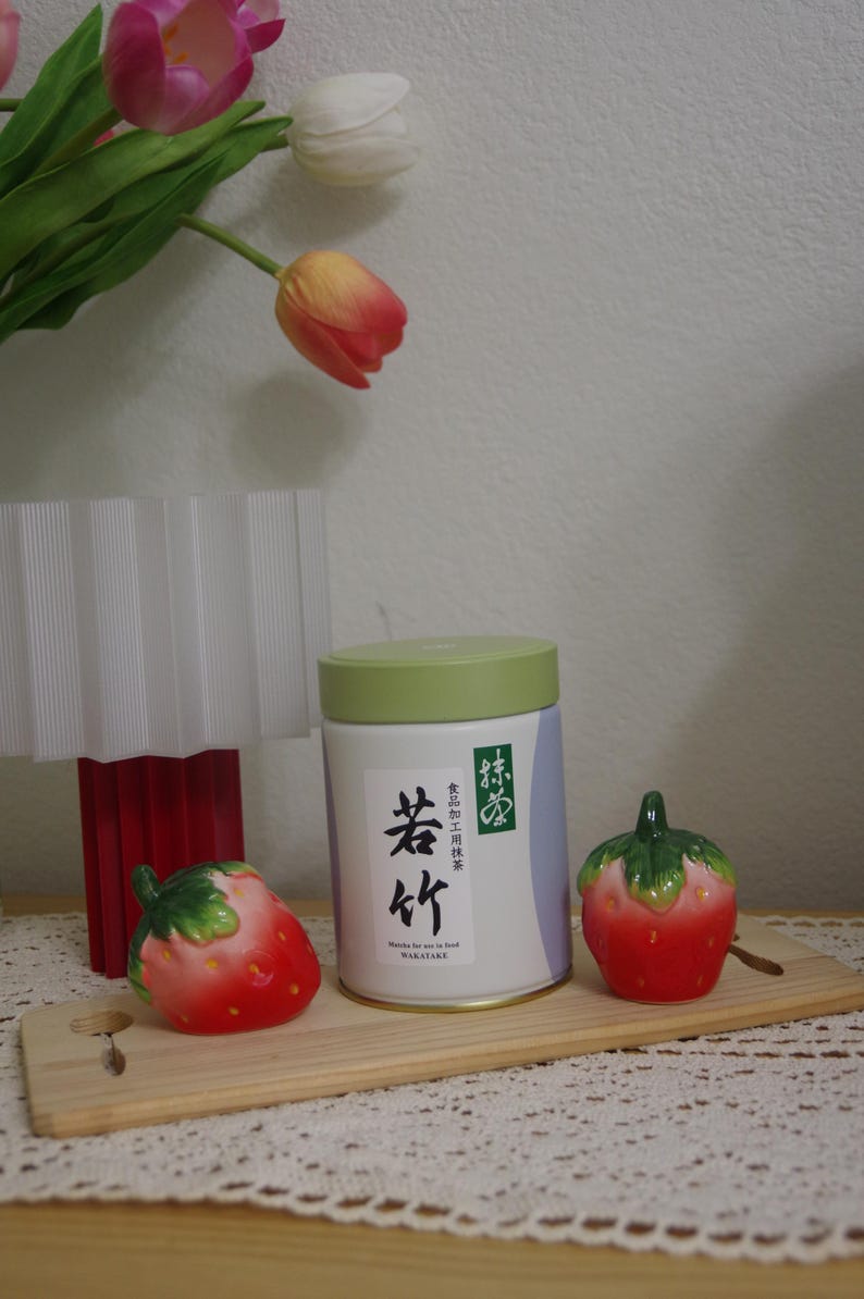 May include: A still-life arrangement featuring a white and green matcha tea tin with Japanese text, flanked by two ceramic strawberry figurines. The scene is set on a wooden board, with tulips and a lamp in the background.