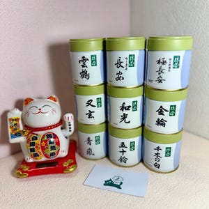 May include: A display of tea canisters with green lids and Japanese text. A white Maneki-neko cat statue with red accents is on the left. A small card with a cat logo is at the bottom.