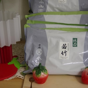 May include: Stack of packaged goods with Japanese text on the front. The packaging is white and gray with green accents. Two red and green strawberry-shaped salt and pepper shakers sit on a wooden cutting board.