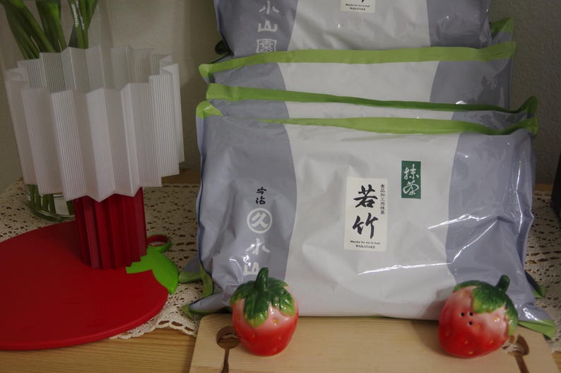 May include: Stack of packaged goods with Japanese text on the front. The packaging is white and gray with green accents. Two red and green strawberry-shaped salt and pepper shakers sit on a wooden cutting board.