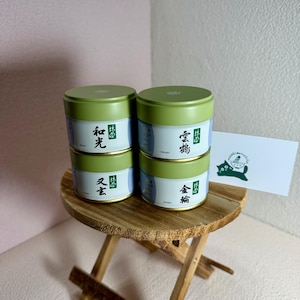 May include: Four small, round, green and white tins stacked on a small wooden table. Each tin has Japanese text and a green lid. A white card with a cat logo is on the table. The tins appear to be tea containers.