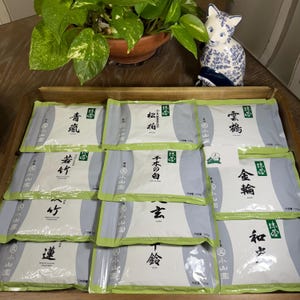 May include: A wooden tray holds several packages of Japanese matcha tea. Each package is white with Japanese text and a green border. A ceramic cat figurine and a potted plant are in the background.