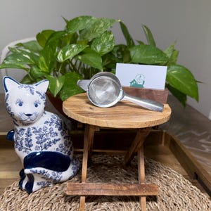 May include: A ceramic cat figurine with blue floral patterns and a stainless steel tea strainer on a small wooden table. A green leafy plant and a business card are in the background. The cat figurine is white with blue accents.