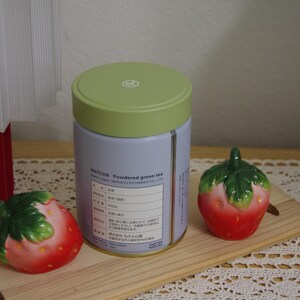 May include: A cylindrical light purple tin of Marukyu Koyamaen matcha powdered green tea with a light green lid, flanked by two ceramic strawberry-shaped shakers on a wooden board. The label displays text including MATCHA.