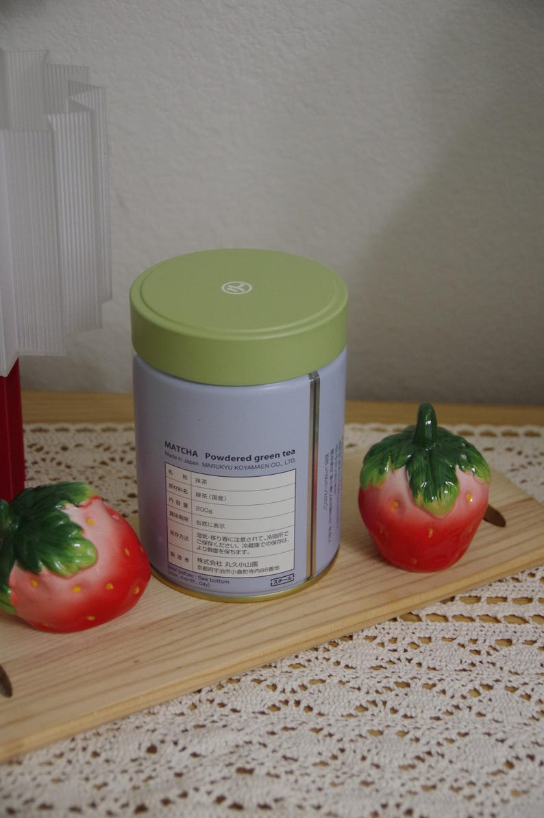 May include: A cylindrical light purple tin of Marukyu Koyamaen matcha powdered green tea with a light green lid, flanked by two ceramic strawberry-shaped shakers on a wooden board. The label displays text including MATCHA.