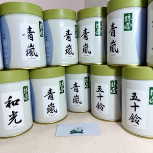 May include: Several cylindrical tea canisters with green lids and white labels are stacked. The labels display Japanese characters and a green logo. Some canisters have a blue wave design. A small card with a green logo is in front.