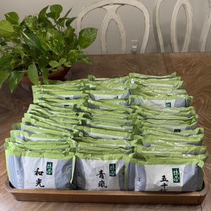 May include: A wooden tray displays many individually wrapped packages. The packages are mainly green and white, with Japanese text and illustrations. A potted plant with green and white speckled leaves is in the background.