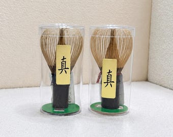 May include: Two bamboo matcha whisks, each enclosed in a clear cylindrical plastic case. The whisks feature light brown tines, a dark brown handle, and a yellow rectangular label with the black Japanese kanji character 真 printed on the front.