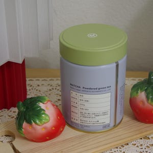 May include: A cylindrical tin of matcha powdered green tea with a light green lid and a light purple label featuring black text, flanked by two ceramic strawberry-shaped shakers with red bodies and green leaves on top.