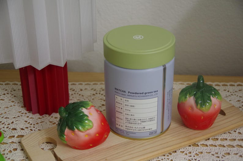May include: A cylindrical tin of matcha powdered green tea with a light green lid and a light purple label featuring black text, flanked by two ceramic strawberry-shaped shakers with red bodies and green leaves on top.