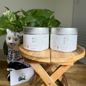 May include: Two silver-lidded tins of organic matcha tea, labelled "Shizuoka" and "Kagoshima" in Japanese and English. The tins are on a small wooden stool, with a ceramic cat figurine and leafy green plant in the background.