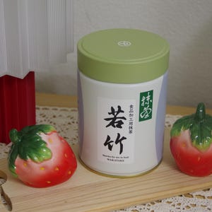 May include: A white and green matcha tea container with Japanese text and two ceramic strawberry figurines on a wooden board. The container has a green lid and a label with black Japanese characters. The strawberries are red with green leaves.