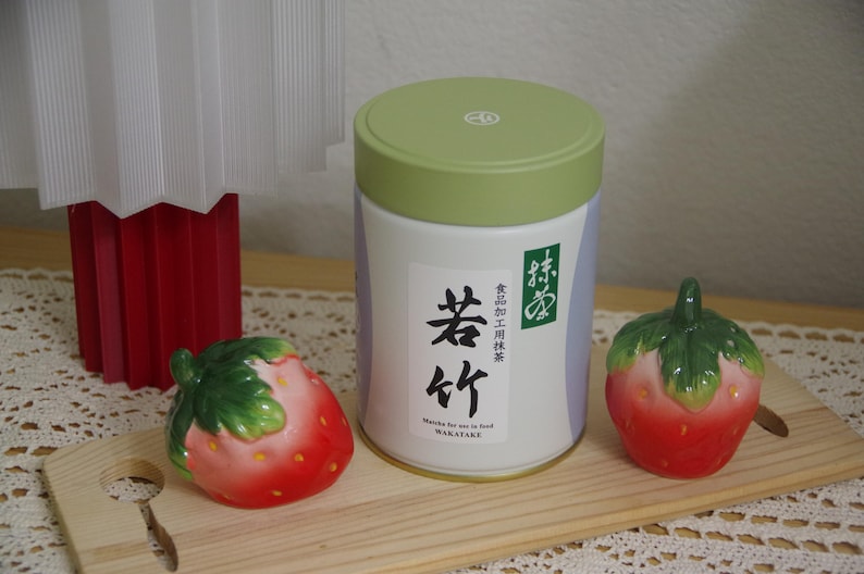 May include: A white and green matcha tea container with Japanese text and two ceramic strawberry figurines on a wooden board. The container has a green lid and a label with black Japanese characters. The strawberries are red with green leaves.