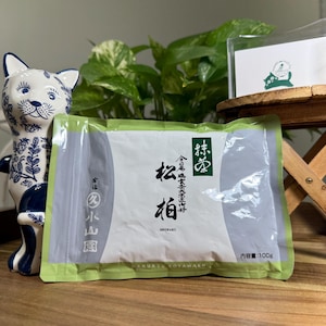 May include: A rectangular green and white pouch of Marukyu Koyamaen Shohaku matcha tea. The label features black Japanese kanji characters, the text SHOHAKU, and the net weight of 100g. A blue and white ceramic cat figurine stands to the left.