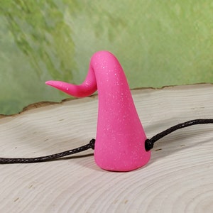 May include: A pair of pink, sparkly devil horns with a black elastic headband. The horns are made of polymer clay and have a slightly curved shape.