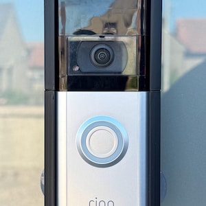 Può includere: Un videocitofono Ring argento e nero montato su una superficie di vetro. Il campanello ha un obiettivo della fotocamera, un pulsante circolare e il marchio "ring" stampato. Progettato per la sicurezza domestica, misura circa 15 cm di altezza.