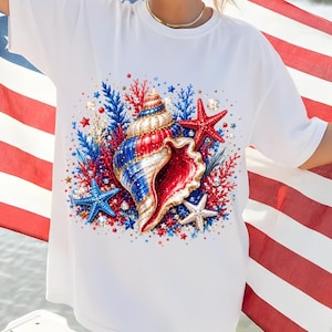 May include: A white short-sleeved t-shirt featuring a central graphic print of a large seashell surrounded by red and blue coral and starfish. The design incorporates a patriotic color palette of red, white, and blue throughout the illustration.