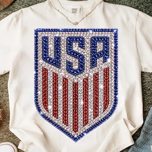 K&ouml;nnte beinhalten: Ein wei&szlig;es T-Shirt mit kurzen &Auml;rmeln und einem gro&szlig;en schildf&ouml;rmigen Aufdruck auf der Brust. Das Motiv besteht aus funkelnden blauen, roten und klaren Strasssteinen, die die Buchstaben USA &uuml;ber vertikalen roten und wei&szlig;en Streifen bilden.