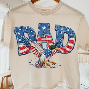Könnte beinhalten: Cremefarbenes T-Shirt mit dem Wort "DAD" in blauen Buchstaben im Denim-Stil mit weißen Sternen. Unter dem Wort ist eine Ente mit rot-weiß-blauen Flügeln abgebildet. Das Shirt hängt an einem Holzbügel.