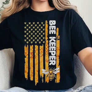 May include: A black t-shirt with a graphic of an American flag with a bee and honeycomb pattern. The text "BEE KEEPER" is printed vertically on the right side of the flag.
