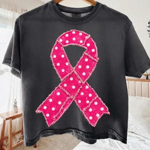 May include: A charcoal grey short-sleeve t-shirt featuring a large, centred awareness ribbon graphic. The ribbon is bright pink with a white polka dot pattern and has a distressed, frayed-edge fabric texture applied to the front of the shirt.