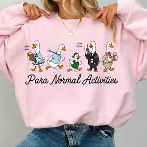 Könnte beinhalten: Hellrosa Sweatshirt mit einem Cartoon-Design von fünf Gänsen in verschiedenen Outfits, darunter ein Anzug und ein Detektiv-Outfit. Der Text "Para Normal Activities" ist unter den Gänsen aufgedruckt.