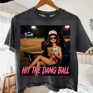 May include: Dark gray t-shirt featuring a graphic of a doll wearing sunglasses and a baseball cap, holding a drink, and sitting in a chair at a baseball field. The text "HIT THE DANG BALL" is printed in pink.