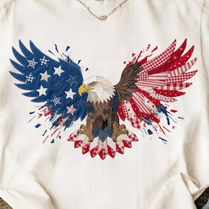 May include: Cream-coloured t-shirt with an eagle motif. The eagle's wings are crafted from patchwork fabrics in red, white, and blue, with star and stripe patterns. The eagle's head has detailed feathers and a yellow beak.