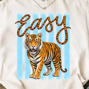 May include: White t-shirt featuring a tiger illustration with orange and black stripes, set against a backdrop of light blue vertical stripes. The word "EASY" is written in an orange, stylized font above the tiger.