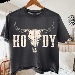 May include: Dark gray t-shirt featuring a white bull skull graphic with the word "HOWDY" in a distressed font. The shirt is hanging on a wooden hanger, showcasing a Western or country theme.