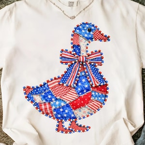 May include: White t-shirt featuring a patriotic goose design. The goose is adorned with a patchwork of red, white, and blue fabrics, including stars and stripes. A beaded bow and trim add detail to the design. The shirt has a v-neck and the brand name is visible.
