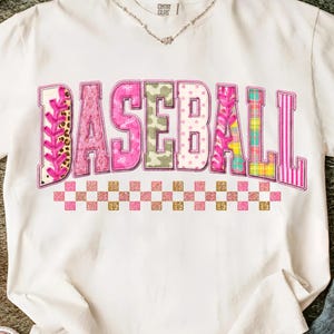 Puede incluir: Camiseta blanca con la palabra "BASEBALL" en letras rosas, verdes y estampadas. La palabra está delineada con purpurina dorada y se encuentra sobre un patrón de cuadros rosas y dorados. Un collar plateado es visible en el escote.