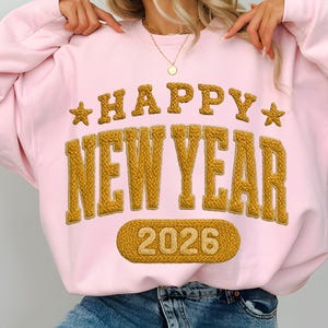 Może przedstawiać: Jasnoróżowa bluza z napisem "HAPPY NEW YEAR 2026" w teksturowanej, złotej czcionce. Bluza ma okrągły dekolt i długie rękawy.