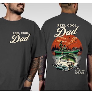 May include: Dark gray t-shirt with the text "REEL COOL Dad" in a cream-colored script on the front. The back features a fishing scene with a boat, two figures, and a large fish, with the text "THE FISHING LEGEND".