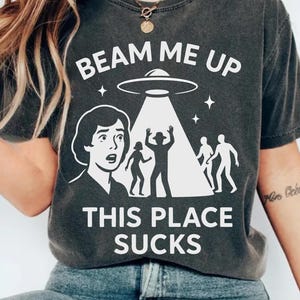 May include: Dark grey t-shirt with the slogan "BEAM ME UP THIS PLACE SUCKS". The design shows a retro-style illustration of a person looking up at a UFO, with figures being beamed up. A casual wear item.
