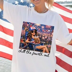 May include: White t-shirt featuring a graphic of a woman relaxing on the Fourth of July, holding a drink. The shirt has the text "chill the fourth out" in a script font. The person is holding an American flag.